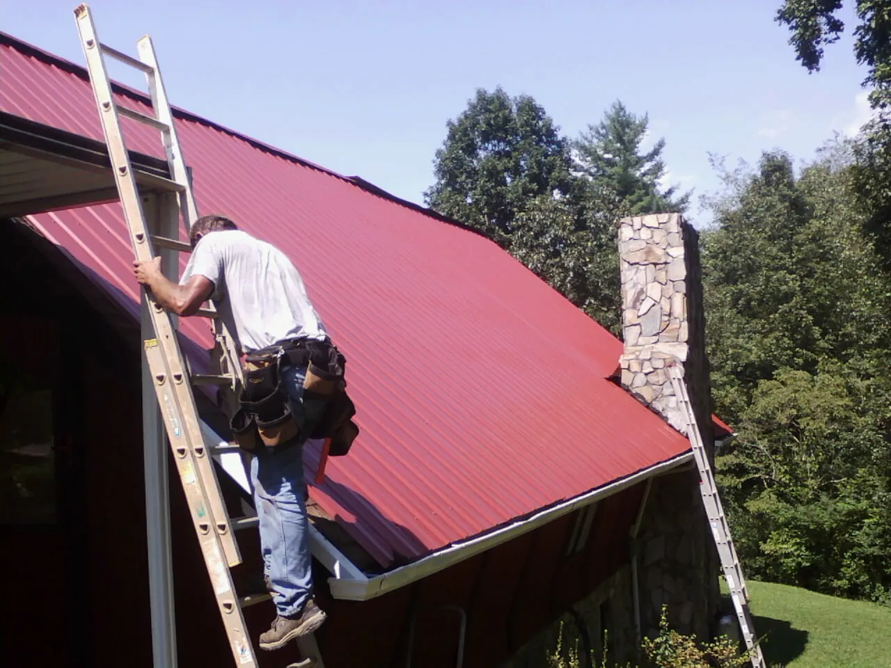 Roofer on a ladder beside a red metal roof and stone chimney — Soffit & Fascia Repair by Roofing Brothers in Greene